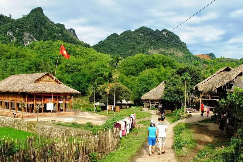 Du khách đi bộ qua những nhà sàn lợp lá và lá cờ Việt Nam giữa bản làng Mai Châu bao quanh núi. Du khách đi bộ qua những nhà sàn lợp lá và lá cờ Việt Nam giữa bản làng Mai Châu bao quanh núi.