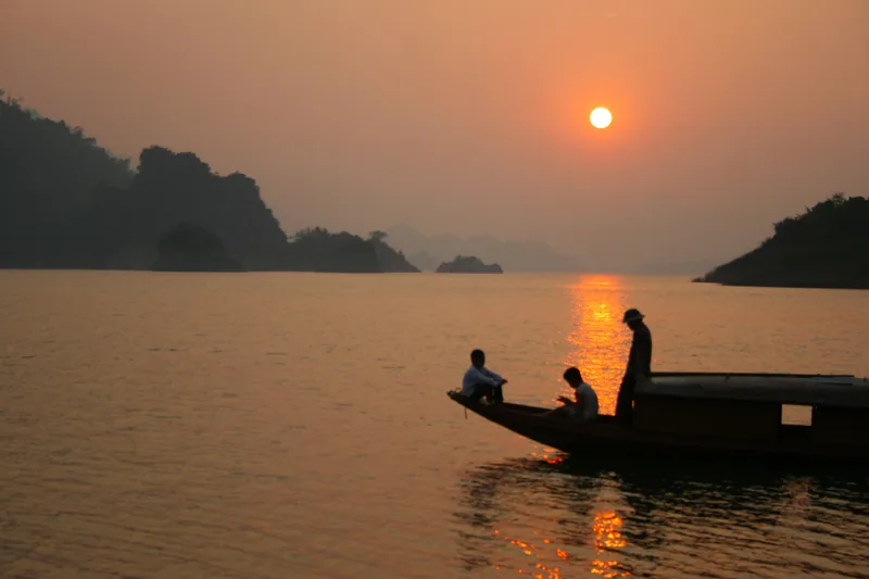 Một chiếc thuyền gỗ chở ba người lướt trên hồ Hòa Bình khi hoàng hôn buông xuống sau những dãy núi mờ sương. Một chiếc thuyền gỗ chở ba người lướt trên hồ Hòa Bình khi hoàng hôn buông xuống sau những dãy núi mờ sương.